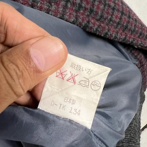 Puede incluir: Primer plano del interior de una prenda, que revela una etiqueta de cuidado con texto y s&iacute;mbolos japoneses. La etiqueta blanca con texto negro est&aacute; adherida a un forro azul. La tela exterior es una mezcla de gris, burdeos y negro.
