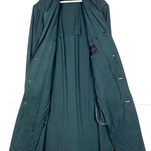 Może przedstawiać: Długi, ciemnozielony płaszcz o klasycznym kroju. Płaszcz posiada wykładany kołnierz, długie rękawy i zapinany na guziki prz&oacute;d. Widoczna jest wewnętrzna podszewka, a płaszcz wisi na białym wieszaku.