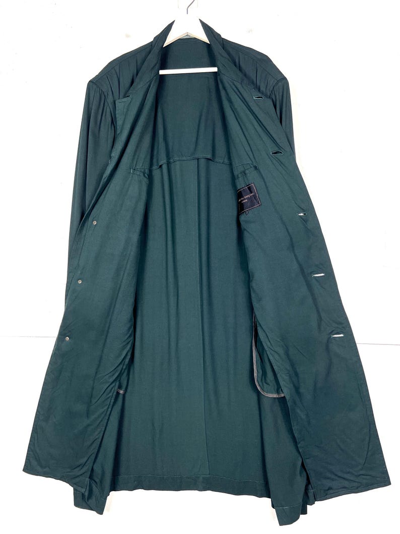 Może przedstawiać: Długi, ciemnozielony płaszcz o klasycznym kroju. Płaszcz posiada wykładany kołnierz, długie rękawy i zapinany na guziki prz&oacute;d. Widoczna jest wewnętrzna podszewka, a płaszcz wisi na białym wieszaku.