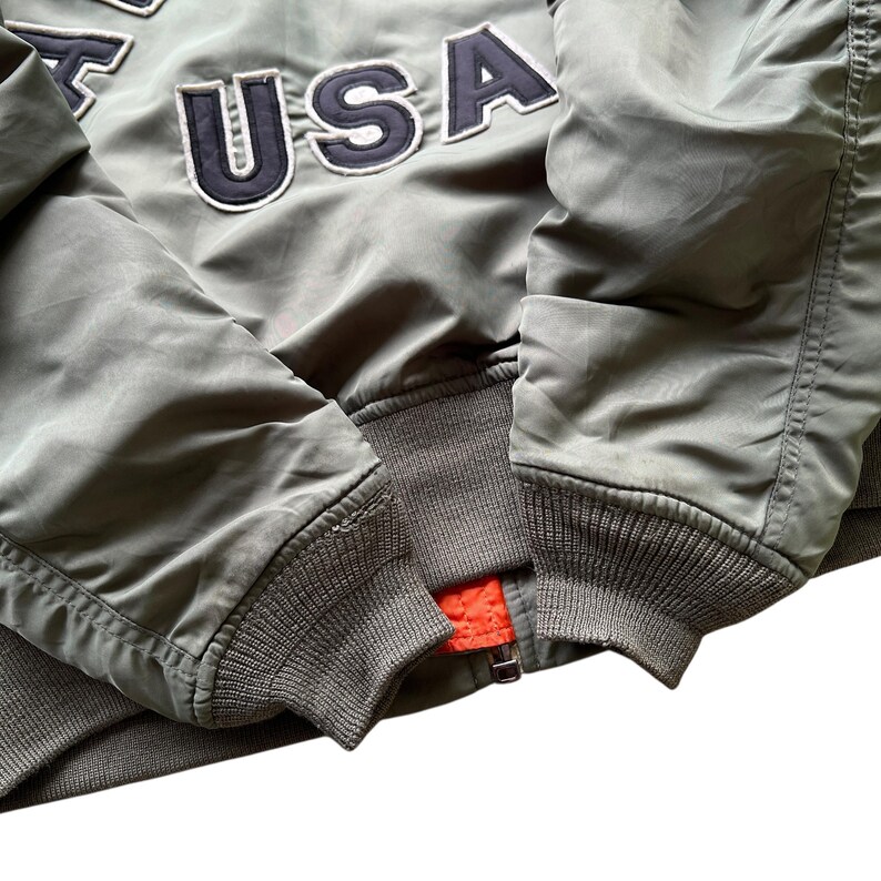 May include: An olive green bomber jacket featuring dark navy blue embroidered letters that spell out USA. The jacket has ribbed knit cuffs and a hem in a matching olive green tone, with a bright orange lining visible near the metal zip closure.