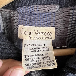Puede incluir: Primer plano de una etiqueta Gianni Versace, Made in Italy. La etiqueta es beige con texto negro, que enumera la composici&oacute;n de la tela: 100% lana y 50% lana, 50% algod&oacute;n. La chaqueta es gris oscuro con forro azul y gris.