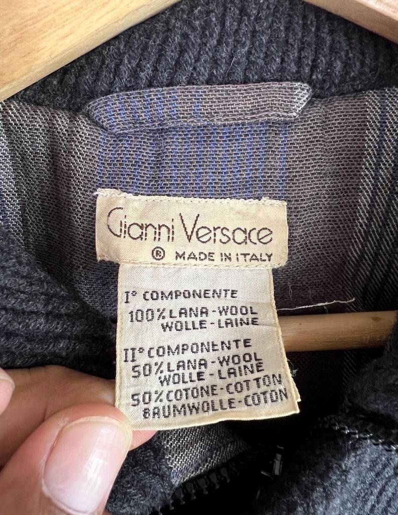Puede incluir: Primer plano de una etiqueta Gianni Versace, Made in Italy. La etiqueta es beige con texto negro, que enumera la composici&oacute;n de la tela: 100% lana y 50% lana, 50% algod&oacute;n. La chaqueta es gris oscuro con forro azul y gris.