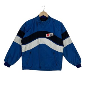 May include: A blue jacket with a high collar and an asymmetrical zipper closure. The front features a horizontal wave pattern in black and white, with a rectangular patch on the chest displaying the red text JS and the black text JAPAN SPEED.