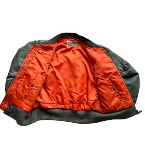 May include: An olive green bomber jacket featuring a bright orange interior lining. The jacket includes a ribbed collar, cuffs, and hem, with two front pockets featuring snap closures and a small utility pocket on the sleeve with a metal zipper.