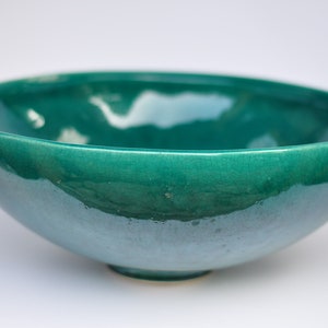 May include: A teal ceramic bowl with a glossy finish. The bowl has a rounded shape with a slightly flared rim and a small, circular base. The surface shows subtle crackling, adding texture to the piece.
