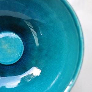 Puede incluir: Un cuenco de cerámica azul turquesa con un acabado brillante. El cuenco tiene una forma circular y un borde ligeramente elevado.
