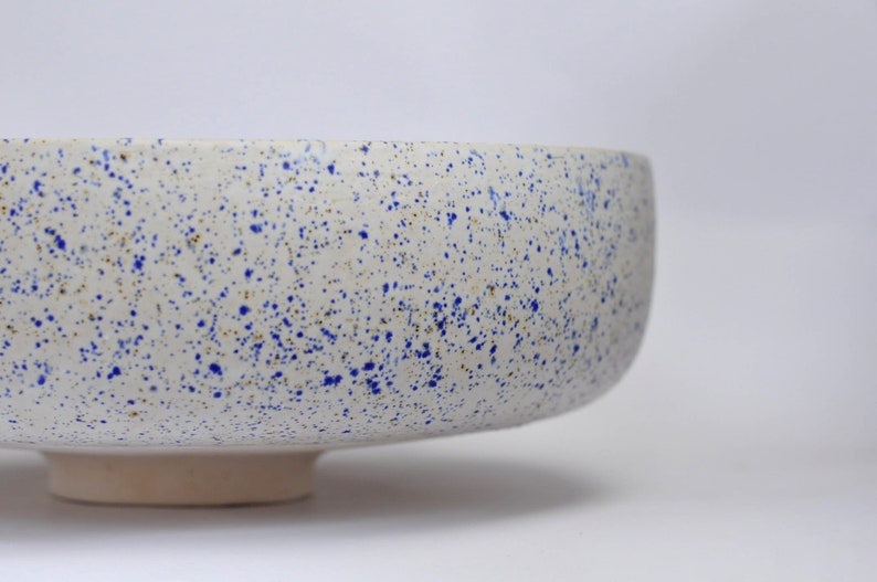 May include: A white ceramic bowl with a speckled blue and brown glaze. The bowl has a wide, rounded base and a slightly flared rim.