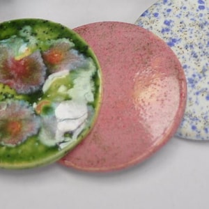 May include: Three round ceramic coasters with different designs. One coaster is green with pink and white floral patterns, another is pink with a speckled texture, and the third is white with blue and gold speckles.