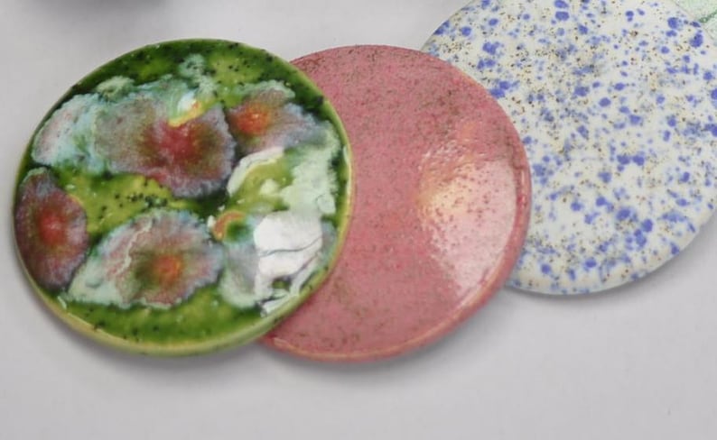 May include: Three round ceramic coasters with different designs. One coaster is green with pink and white floral patterns, another is pink with a speckled texture, and the third is white with blue and gold speckles.