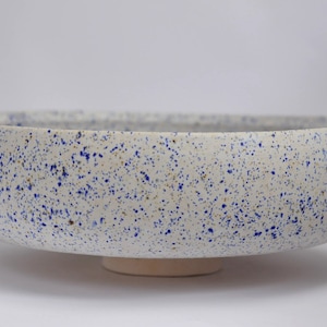 May include: A large white ceramic bowl with a speckled blue and white glaze. The bowl has a wide, shallow base and a rounded rim.