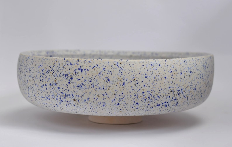 May include: A large white ceramic bowl with a speckled blue and white glaze. The bowl has a wide, shallow base and a rounded rim.