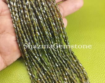 May include: A collection of olive-green gemstone beads. The beads are small, oblong, and faceted, creating a sparkling effect. The text "ShazmaGemstone" is visible in the image. The beads are held against a green background.