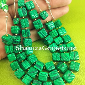 Puede incluir: Primer plano de un collar hecho de cuentas cuadradas de malaquita verde. Las cuentas tienen un patrón de remolino verde oscuro y están separadas por espaciadores dorados. El texto "Shamza Gemstone" es visible.