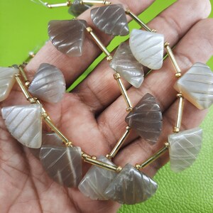 May include: A strand of gray and white sunstone beads with a gold spacer between each bead. The beads are carved in a fan shape.