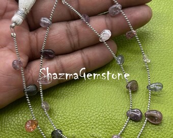 May include: Strands of gemstone beads in various colors, including clear, purple, orange, and gray. The beads are strung on a silver wire and are teardrop-shaped. The image includes white tassels and the text "ShazmaGemstone".