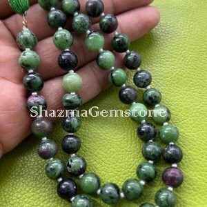 May include: A close-up of a beaded necklace featuring green and black gemstones with a few red-purple stones. The necklace has a green tassel and the text "Shazma Gemstone". The beads are strung on a white string.