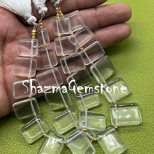 Puede incluir: Cuentas de piedras preciosas rectangulares transparentes ensartadas, con detalles dorados. Las cuentas se muestran con borlas blancas. El texto "Shazma Gemstone" es visible.