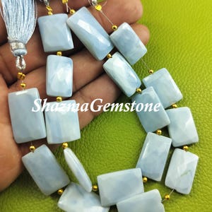 Puede incluir: Una hebra de cuentas de piedra preciosa rectangulares de color azul claro con separadores dorados. Las cuentas están enhebradas en un alambre delgado y tienen un acabado liso y pulido.  ShazmaGemstone está impreso en la imagen.