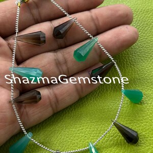 Könnte beinhalten: Stränge von tropfenförmigen Edelsteinperlen in Grün-, Braun- und Türkistönen, auf einer silbernen Kette aufgereiht. Die facettierten Perlen sind vor einem grünen Hintergrund zu sehen. Oben ist eine bunte Quaste zu sehen. Der Text "ShazmaGemstone" ist auf dem Bild aufgedruckt.