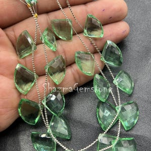 May include: Two strands of faceted, light green gemstones with a leaf-like shape. The gemstones are strung on silver-colored wire and are accented with green tassels. The text "shazmaGemstone" is visible.