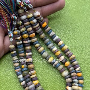 May include: Three strands of multicolored gemstone beads in shades of gray, brown, yellow, and purple, shaped like small flattened discs. Each strand is finished with a vibrant, multi-colored silk tassel at the top. The text ShazmaGemstone is visible.