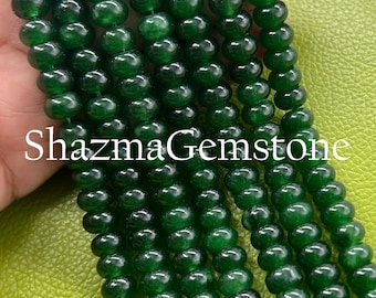 May include: Close-up of strands of glossy, deep green gemstone beads. The beads are round and polished. The text "ShazmaGemstone" is visible in white. The beads are held in a hand against a green background.