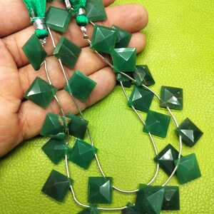 May include: A close-up of green gemstone beads. The beads are square-shaped with faceted edges, strung on silver wire. The image also shows green tassels. The background is a solid, bright green.