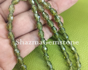 May include: Olive green faceted gemstone beads strung together. The beads are oval-shaped and have a sparkling, reflective surface. The text "Shazma Gemstone" is visible in the image. The beads are on a green string.