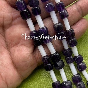 May include: Close-up of strands of amethyst gemstone beads. The beads are a deep purple colour and are interspersed with white cylindrical spacers. The text "ShazmaGemstone" is visible on the image.