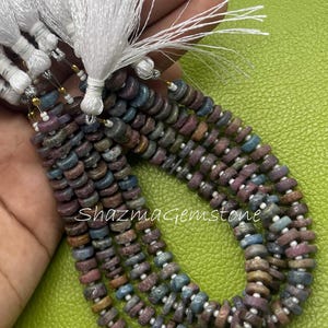 May include: Multiple strands of small, disc-shaped multicoloured gemstone beads in shades of blue, purple, and brown, interspersed with tiny white spacer beads. Each strand is finished with a white tassel. The text shazmagemstone is visible.