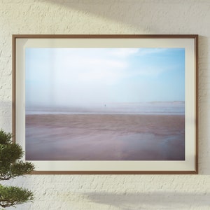 Op de afbeelding: Een ingelijste foto van een strandsc&egrave;ne met een enkele figuur die in de verte loopt. De lucht is lichtblauw met wat wolken en het zand is lichtbruin. De foto is ingelijst in een eenvoudig bruin frame.