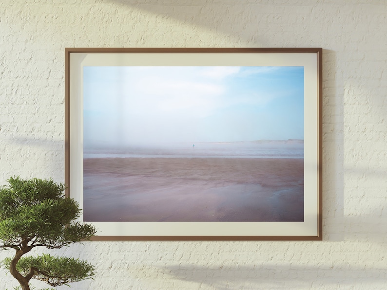 Op de afbeelding: Een ingelijste foto van een strandsc&egrave;ne met een enkele figuur die in de verte loopt. De lucht is lichtblauw met wat wolken en het zand is lichtbruin. De foto is ingelijst in een eenvoudig bruin frame.