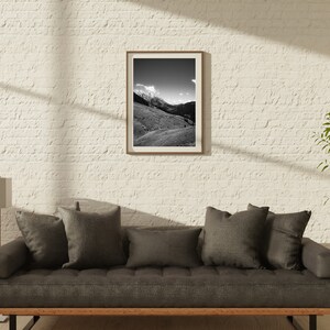 Op de afbeelding: Een grijze bank met donkergrijze kussens staat voor een witte bakstenen muur. Een zwart-witte ingelijste foto van een berglandschap hangt aan de muur. Een groene plant staat in een pot op een kleine tafel rechts van de bank.