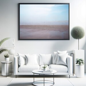 Op de afbeelding: Een witte bank met witte kussens staat voor een ingelijst foto van een strandscene. Een ronde salontafel met een zwart metalen frame staat voor de bank. Een kleine plant in een pot staat op de salontafel. Een wit tapijt bedekt de vloer.