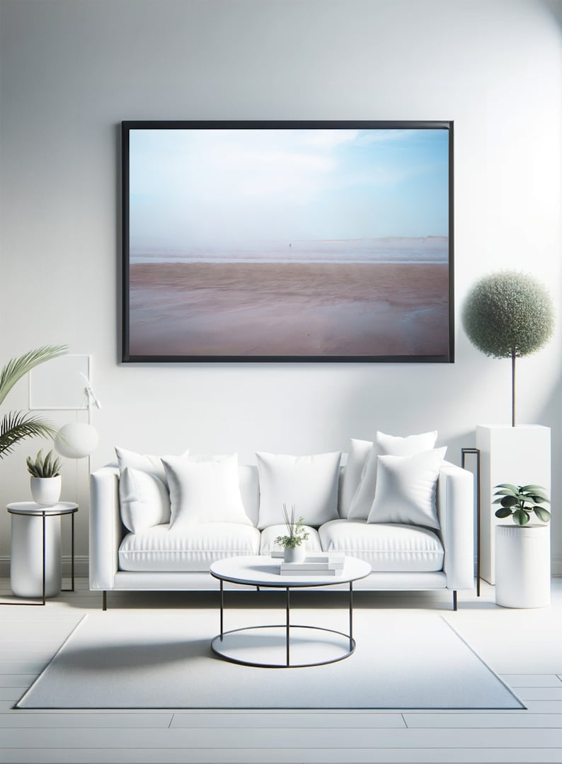 Op de afbeelding: Een witte bank met witte kussens staat voor een ingelijst foto van een strandscene. Een ronde salontafel met een zwart metalen frame staat voor de bank. Een kleine plant in een pot staat op de salontafel. Een wit tapijt bedekt de vloer.