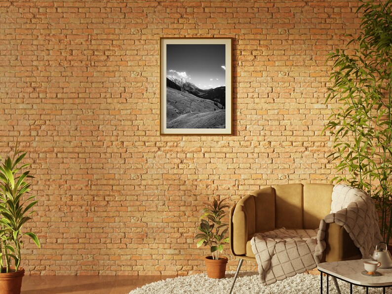 Op de afbeelding: Een bruine fauteuil met een wit en bruin plaid staat in een kamer met een bakstenen muur. Een zwart-witte ingelijste foto van een berglandschap hangt aan de muur. Een kleine plant in een pot staat op de vloer voor de fauteuil.