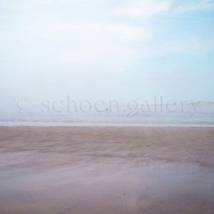 Op de afbeelding: Een mistige strandscene met een enkele figuur die in de verte loopt. Het zand is lichtbruin en de lucht is lichtblauw.
