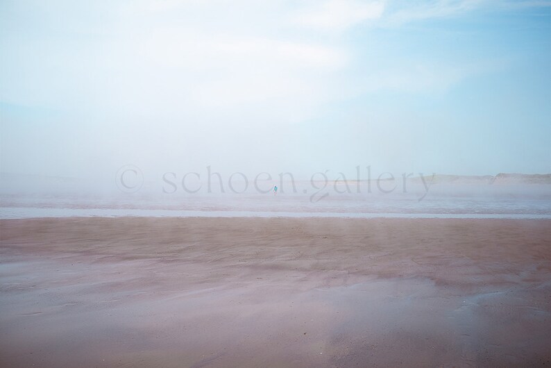Op de afbeelding: Een mistige strandscene met een enkele figuur die in de verte loopt. Het zand is lichtbruin en de lucht is lichtblauw.