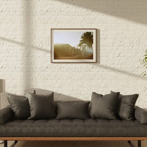Op de afbeelding: Een grijze bank met donkergrijze kussens in een woonkamer met een witte bakstenen muur. Een ingelijste foto van een woestijnlandschap hangt aan de muur boven de bank. Een hoge, gouden staande lamp staat links van de bank. Een plant in een pot staat rechts van de bank.