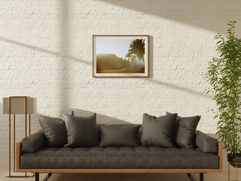 Op de afbeelding: Een grijze bank met donkergrijze kussens in een woonkamer met een witte bakstenen muur. Een ingelijste foto van een woestijnlandschap hangt aan de muur boven de bank. Een hoge, gouden staande lamp staat links van de bank. Een plant in een pot staat rechts van de bank.