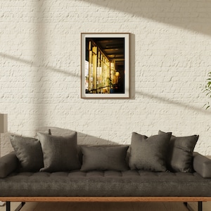 Puede incluir: Una fotograf&iacute;a enmarcada de un edificio con grandes ventanales, mostrando un interior luminoso con una l&aacute;mpara y plantas. La fotograf&iacute;a est&aacute; colgada en una pared de ladrillo blanco sobre un sof&aacute; gris con cojines.