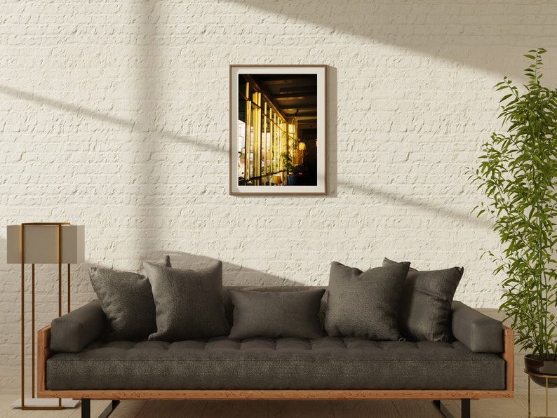 Puede incluir: Una fotograf&iacute;a enmarcada de un edificio con grandes ventanales, mostrando un interior luminoso con una l&aacute;mpara y plantas. La fotograf&iacute;a est&aacute; colgada en una pared de ladrillo blanco sobre un sof&aacute; gris con cojines.