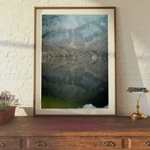 May include: A framed photograph of a mountain lake with a reflection of the surrounding trees and mountains. The water is a deep green color and the sky is a light blue.