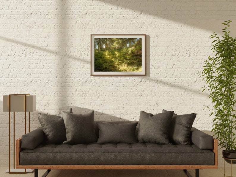 May include: A gray sofa with dark gray cushions sits in a room with a white brick wall. A framed photograph of a forest scene hangs on the wall above the sofa. A tall gold floor lamp stands to the left of the sofa. A potted plant is in the corner of the room.