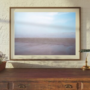 Op de afbeelding: Een ingelijste foto van een strandscene met een enkele persoon die in de verte loopt. Het strand is zanderig en het water is lichtblauw. De lucht is lichtblauw met wat wolken. De lijst is lichtbruin.