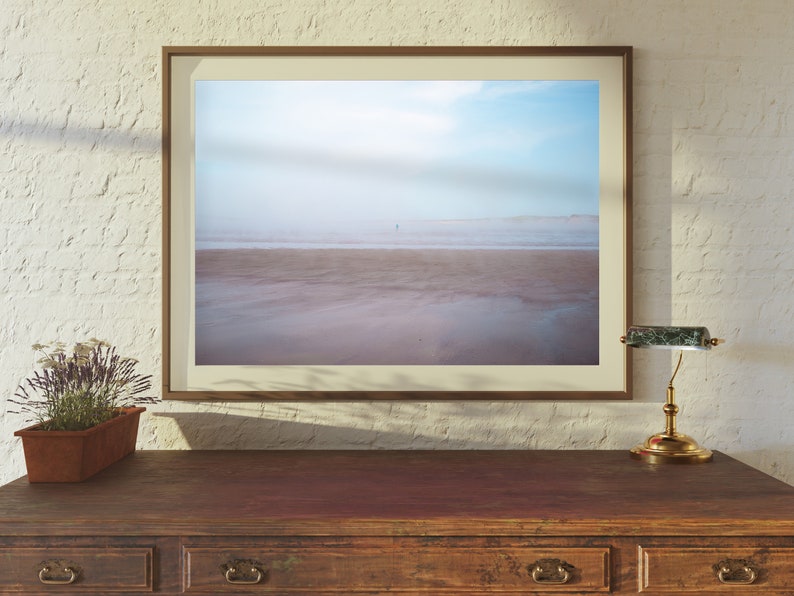 Op de afbeelding: Een ingelijste foto van een strandscene met een enkele persoon die in de verte loopt. Het strand is zanderig en het water is lichtblauw. De lucht is lichtblauw met wat wolken. De lijst is lichtbruin.