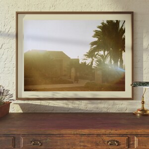 Op de afbeelding: Een ingelijste foto van een gebouw met palmbomen op de achtergrond. De foto is in een bruin frame en hangt aan een witte muur. Op de voorgrond staat een klein tafeltje met een lamp.
