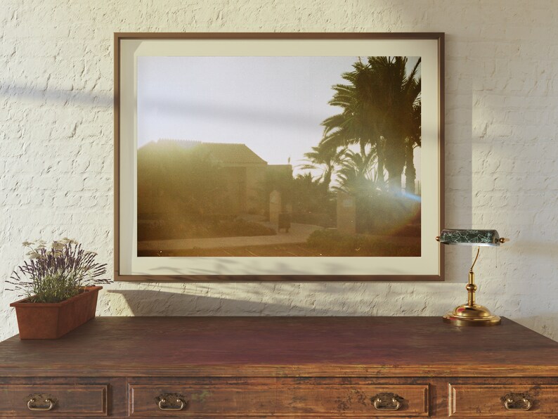 Op de afbeelding: Een ingelijste foto van een gebouw met palmbomen op de achtergrond. De foto is in een bruin frame en hangt aan een witte muur. Op de voorgrond staat een klein tafeltje met een lamp.
