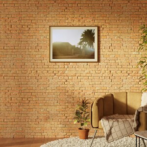 Op de afbeelding: Een bruine fauteuil met een wit plaid staat voor een bakstenen muur. Een ingelijste foto van een gebouw met palmbomen hangt aan de muur. Een kleine plant in een pot staat op de vloer voor de fauteuil. Een kleine tafel met een kopje en schoteltje staat naast de fauteuil.