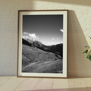 Op de afbeelding: Een zwart-witfoto van een bergketen met een bewolkte lucht. De foto is ingelijst in een bruin houten lijst en hangt aan een witte bakstenen muur.